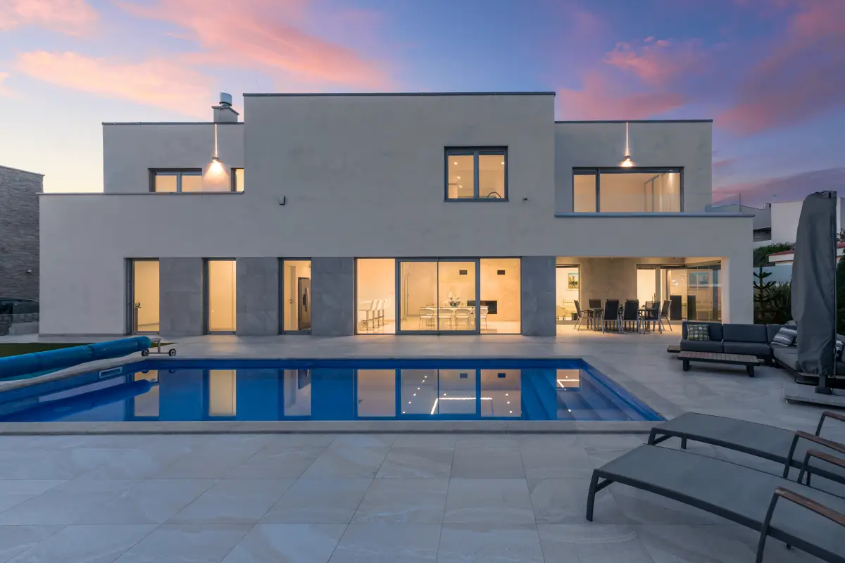 Fotografija luksuzne počitniške vile z osvetljenim bazenom ob mraku, ki ustvarja idilično vzdušje in močno željo po nepozabnem dopustu ter rezervaciji.